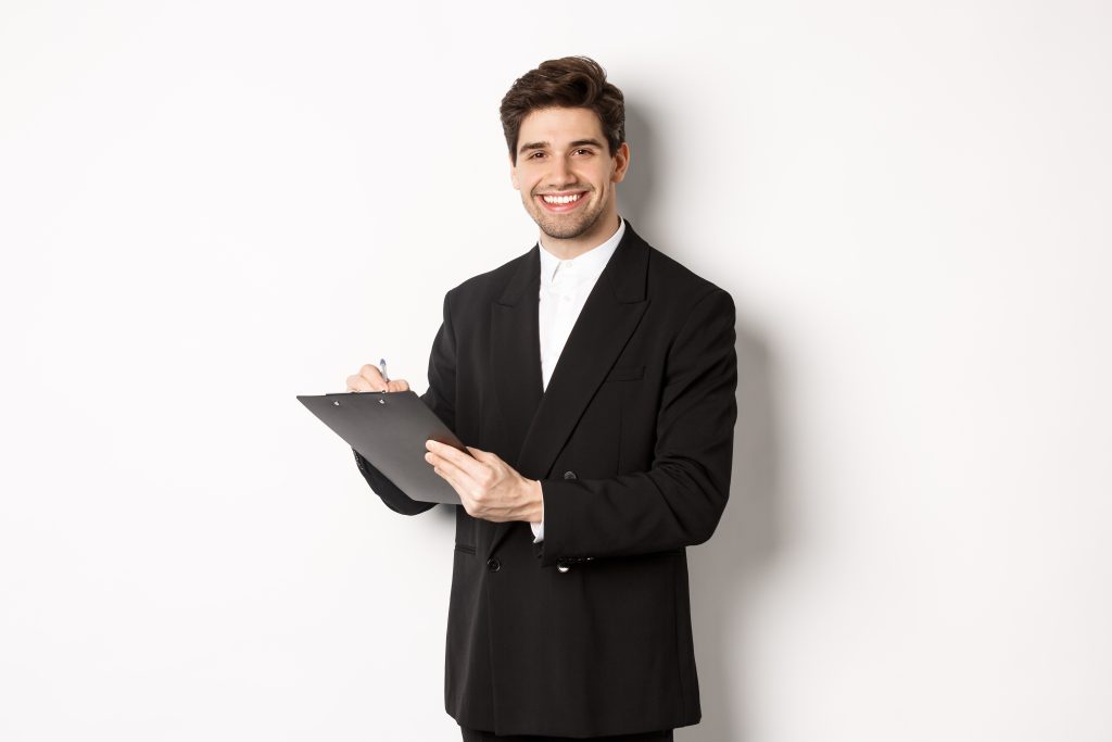 Portrait of confident businessman in black suit, signing documents and smiling, standing happy against white background